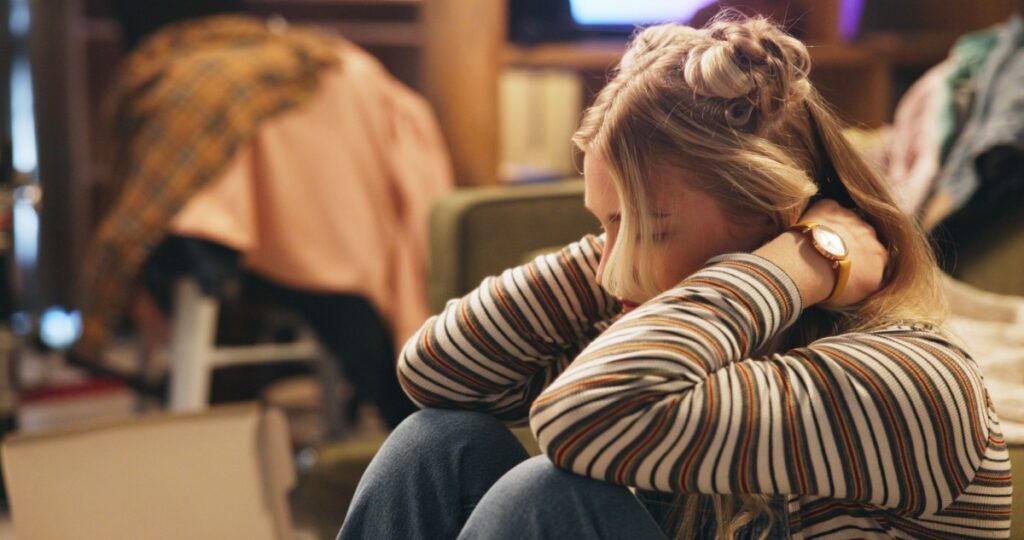 a depressed college student in her dorm room, holding her head with piles of clothes everywhere from not being able to clean