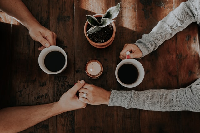 an overhead shot of a couple holding hands and coffee mugs across a wood table