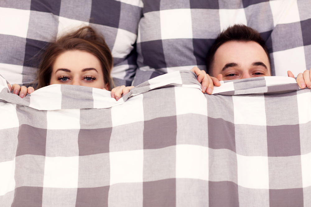 Adult male and female couple peeking out cheekily from a blanket on their bed
